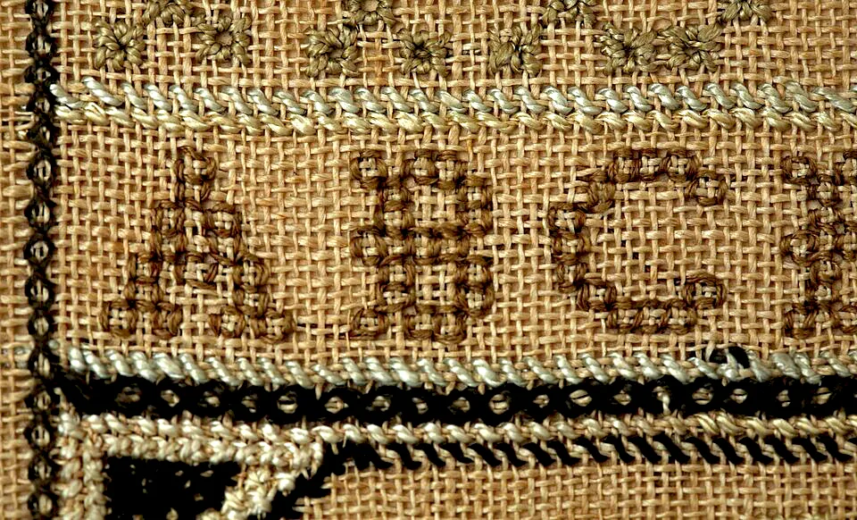 Close-up detail of an antique needlework sampler by Mary Keeler, showing cross stitch characters on linen with counted backstitch linework and a floral border.