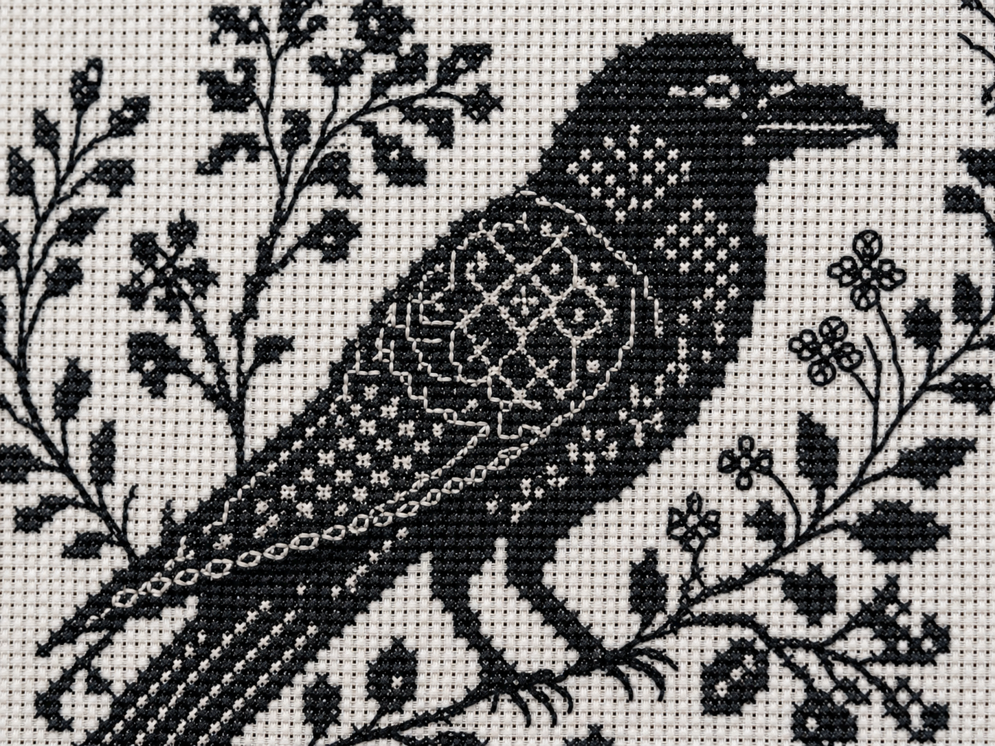 Stitched blackwork cross stitch piece of a crow on white Aida, with geometric repeated fills shading the body and delicate botanical linework framing the bird