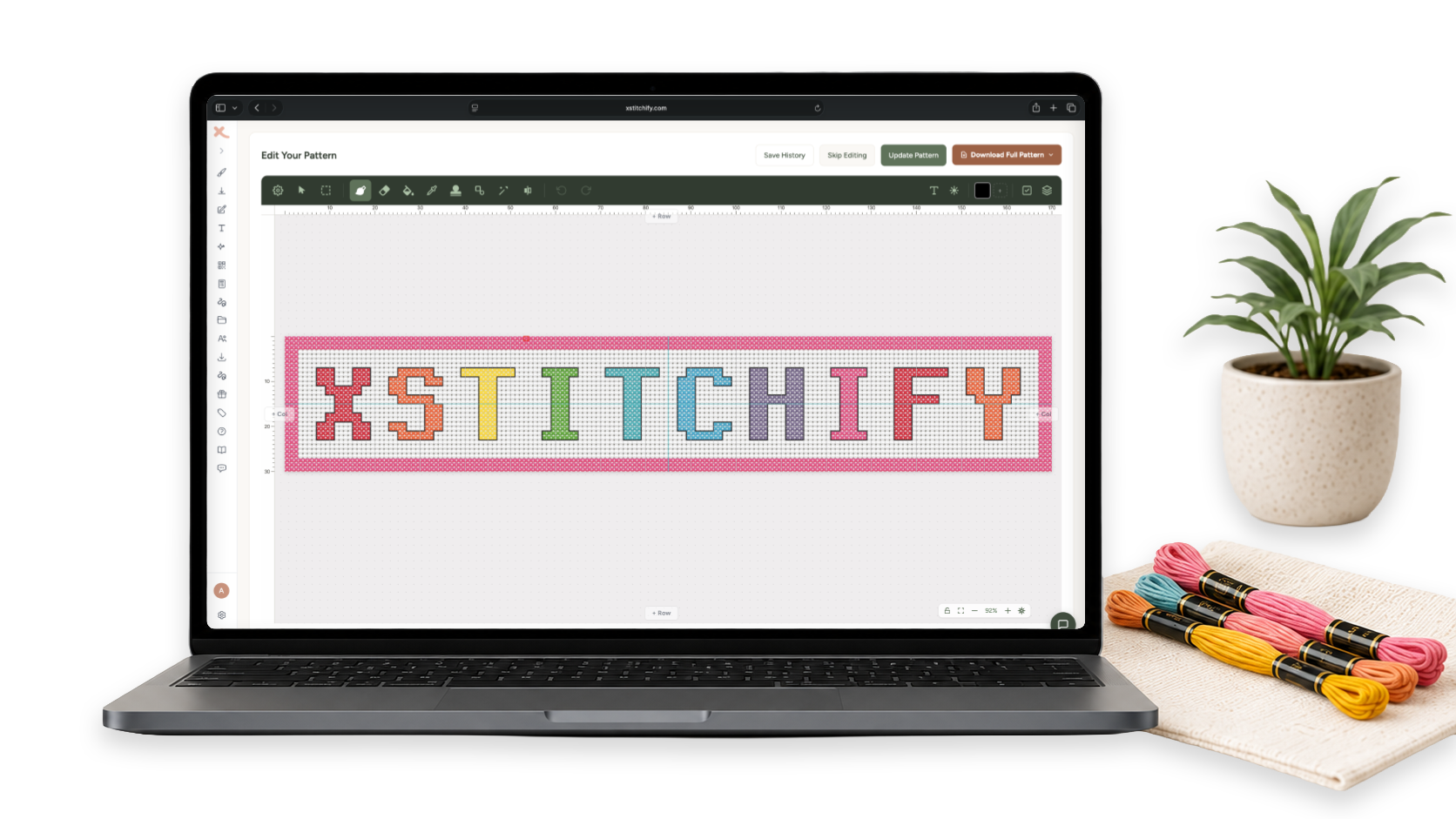 Xstitchify pattern editor on a laptop showing a colourful XSTITCHIFY cross stitch text pattern, beside embroidery floss and a plant